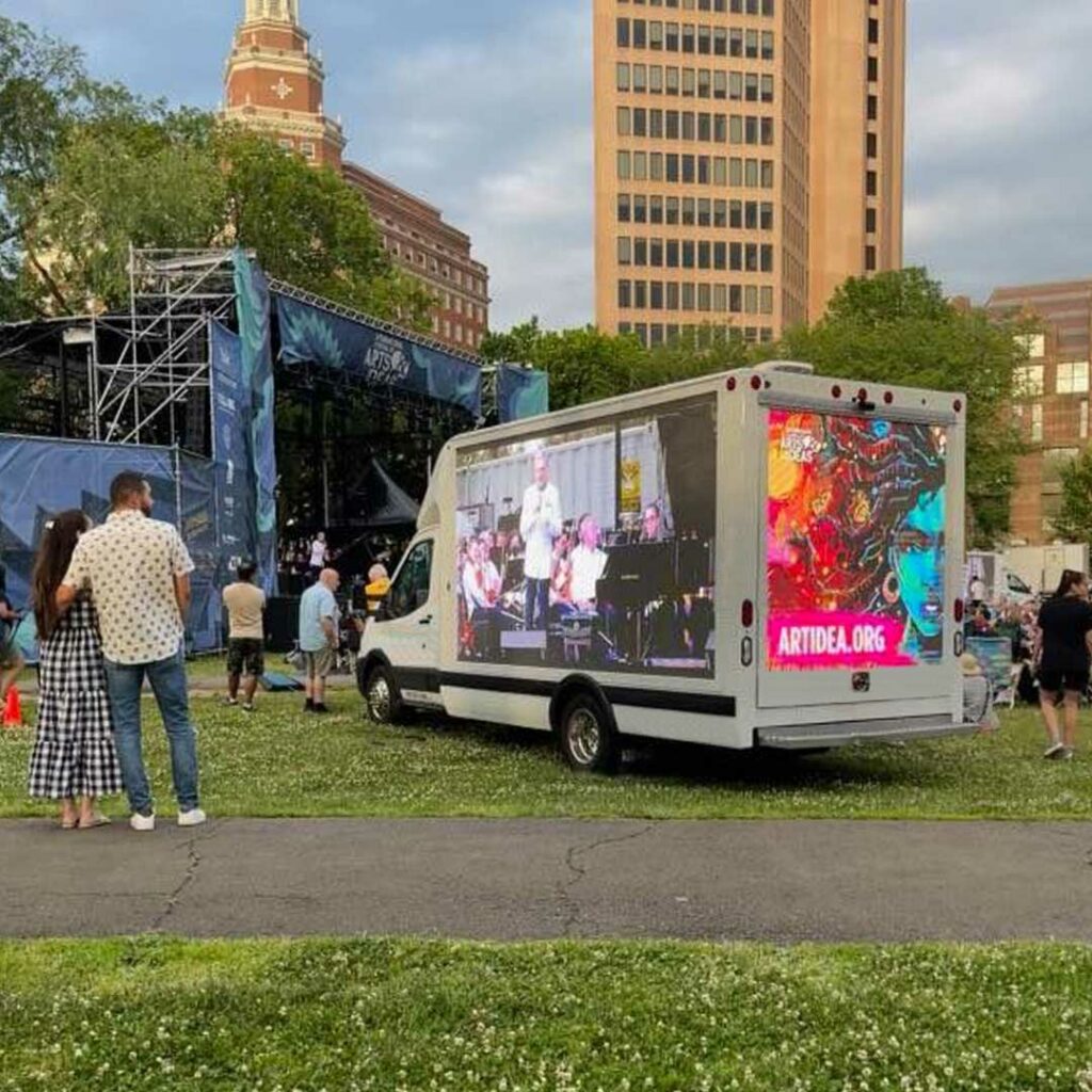 The BrandVan parked at a concert, livestreaming on the sides for a closer look for guests further back while advertising for sponsors on the back screen.